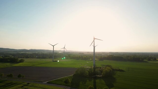 Large wind turbines with blades in field aerial view bright orange sunset blue sky wind park slow motion drone turn. Silhouettes windmills, large orange sun disc summer lens flare. Alternative energy