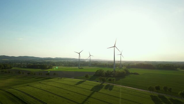 Large wind turbines with blades in field aerial view bright orange sunset blue sky wind park slow motion drone turn. Silhouettes windmills, large orange sun disc summer lens flare. Alternative energy
