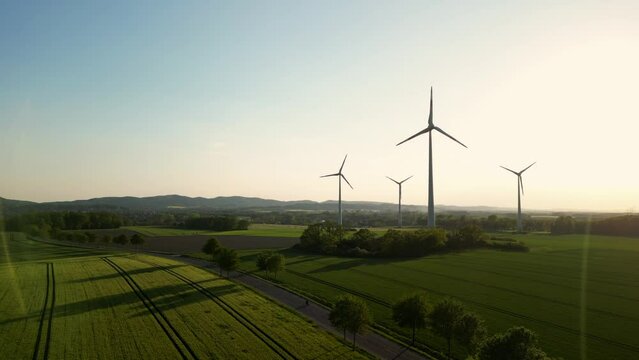 Large wind turbines with blades in field aerial view bright orange sunset blue sky wind park slow motion drone turn. Silhouettes windmills, large orange sun disc summer lens flare. Alternative energy