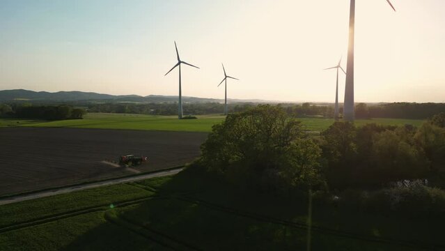Large wind turbines with blades in field aerial, a tractor watering a sown field. Silhouettes windmills, large orange sun disc summer lens flare. Alternative energy,