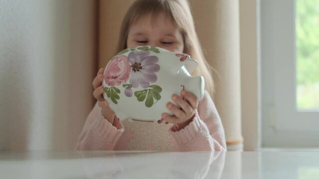 The child shakes the piggy bank and listens to the clinking of coins, and then turns it over and tries to shake it