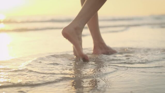Slim female legs and feet walking along sea water waves on sandy beach. Pretty woman walks at seaside surf. Splashes of water and foam, with the sunset reflecting on the water surface.