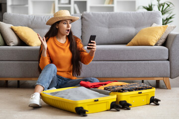 Shocked young woman traveller looking at phone screen, home interior