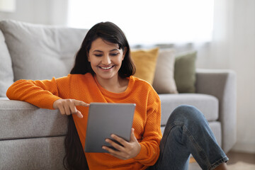 Happy eastern woman sitting on floor at home, using tablet