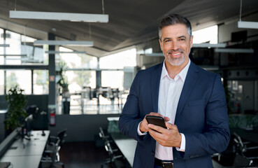 Happy mid aged business man ceo standing in office using mobile phone. Mature businessman professional executive manager digital crypto investor holding smartphone satisfied with corporate solution. - obrazy, fototapety, plakaty