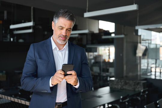 Busy mid aged business man ceo wearing blue suit standing in office using mobile cell phone. Mature businessman professional executive manager holding smartphone checking financial apps on cellphone.