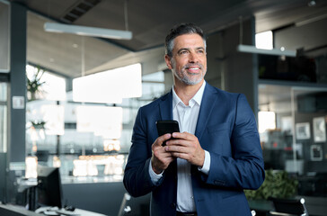 Naklejka na ściany i meble Smiling happy confident mid aged male company ceo executive wearing suit holding cellphone standing in office using business mobile apps technology financial online solutions on cell phone.