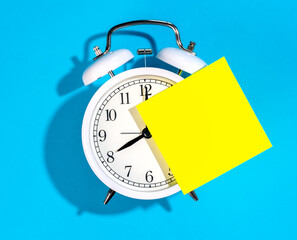 White alarm clock and blank yellow paper sticker on a blue background isolated.