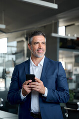 Confident mid aged male company ceo, happy business man executive wearing suit holding cellphone standing in office thinking using mobile apps tech financial online solutions on cell phone. Vertical