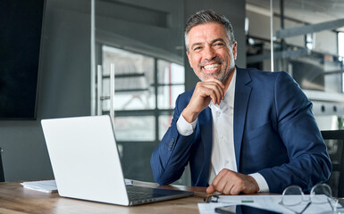 Happy smiling middle aged professional business man company executive ceo manager wearing blue suit sitting at desk in office working on laptop computer laughing at workplace. Portrait. - obrazy, fototapety, plakaty