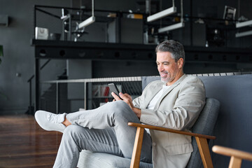 Happy mid aged business man sitting on chair in modern office space using cell phone services and...