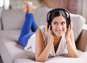 Happy woman, headphones and listening to music on sofa for thinking, comfort or relaxing at home. Female lying on living room couch with headset for audio sound track or relax on holiday break