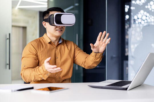 Successful businessman in online meeting uses vr glasses virtual reality, man communicates remotely with partners in metaverse, works inside office sitting at workplace with laptop.