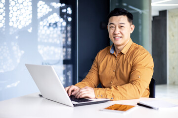 Young and successful asian businessman in working on laptop inside modern office building, happy manager smiling and looking at camera, entrepreneur investor portrait