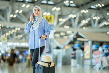 Fototapeta na wymiar An Asian Muslim wearing a blue hijab is preparing for a vacation and she is at the airport. She is using her mobile phone to contact her friends and Muslim travelers.