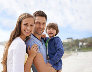 Obraz premium Family, child and portrait outdoor at beach for travel, adventure or holiday in summer with a smile. A man, woman and kid or son together on vacation at sea with a blue sky, parents and happiness