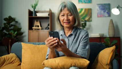 Beautiful elderly woman smile using mobile phone enjoy playing social media checking email. Happy attractive grandma hand holding smartphone scrolling looking at screen sit on sofa in living room