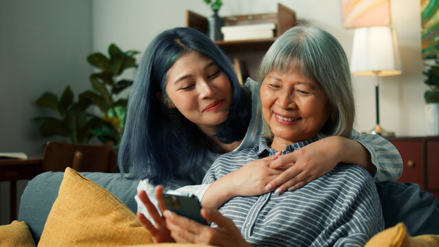 Beautiful Asian elderly woman charmingly smile using smartphone smiling while daughter walk into camera hug mother talk to mom looking at mobile phone screen  Senior Lifestyle Concept