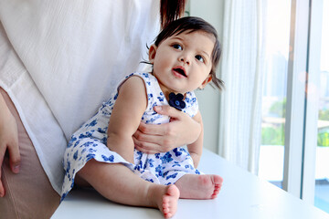 Portrait of happy smiling toddle baby sitting next to window in embrace of her mother parent, little child girl looking at view, mom holding and hugging cute daughter kid, love in family.