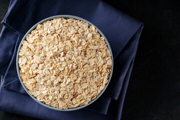 Raw oatmeal in bowl on a blue linen napkin. Oat flakes on a dark background. Healthy eating concept. Copy space