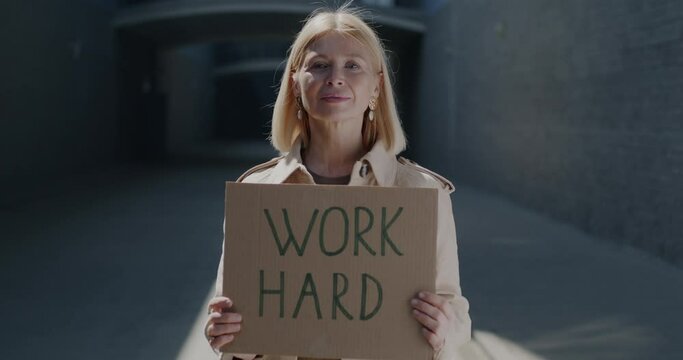 Slow motion portrait of mature lady holding inspirational sign Work Hard standing against urban background. Career and motivation concept.