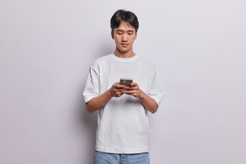 Horizontal shot of handsome Asian man types something on smartphone chats in messenger checks social networks has serious expression dressed in casual t shirt isolated over white background.
