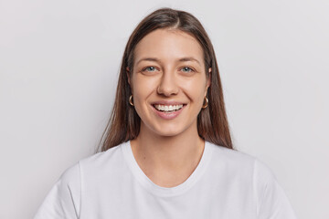 Portrait of good looking woman with dark straight hair smiles broadly expresses positive emotions wears casual t shirt isolated over white wall. European female model poses in studio for making photo