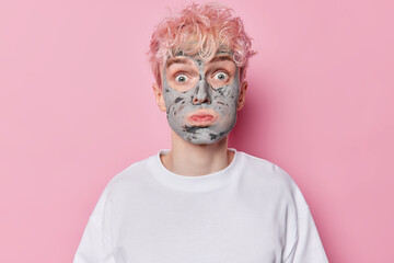 Photo of pink haired young man blows cheeks applies facial clay mask to improve skin texture and refine pores has widely opened eyes dressed in casual white t shirt isolated over rosy background.