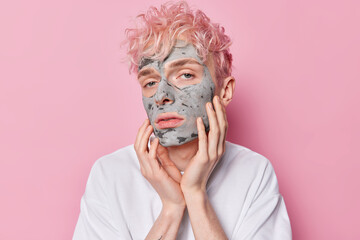 Photo of serious teenage male model applies facial clay mask to improve elasticity of skin removes excess dirt and impurities looks attentively at camera wears white t shirt poses against pink wall