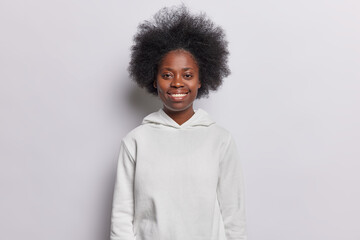 Fototapeta na wymiar Waist up shot of cheerful dark skinned woman smiles pleasantly looks directly at camera wears casual hoodie stands delighted against white background expresses positive emotions enjoys her life