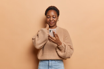 Pretty dark skinned woman keeps hand on neck focused with interest at smartphone smile gently dressed in knitted jumper and jeans isolated over brown background. People communication and technology