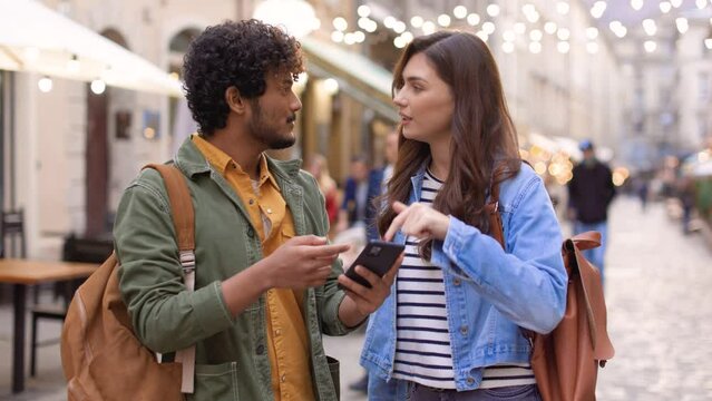 Portrait of joyful couple man and woman looking at smartphone screen scrolling social media standing outdoor in city. Mixed-race handsome guy and beautiful female friends talking on street, urban life