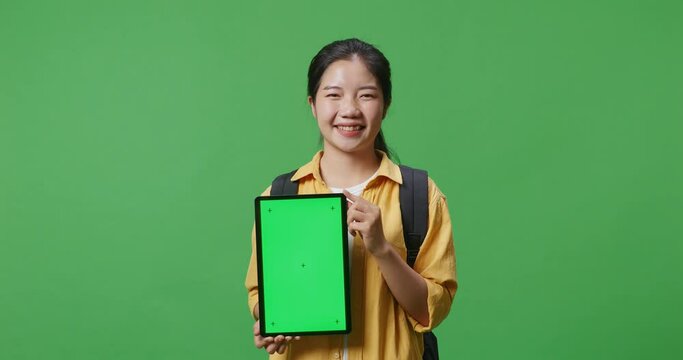 Close Up Of Asian Woman Student With A Backpack Smiling And Showing Green Screen Tablet To Camera While Standing In The Green Screen Background Studio
