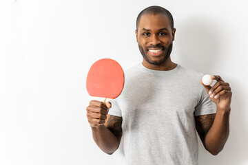 A dark-skinned man is posing on a white background with a ping-pong racket and a ball. The boy shows the team he plays with. Concept of pingpong in African men - obrazy, fototapety, plakaty