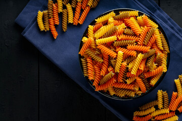 Raw fusilli pasta in bowl on dark wooden background. Tricolor fusilli pasta on a blue linen napkin....