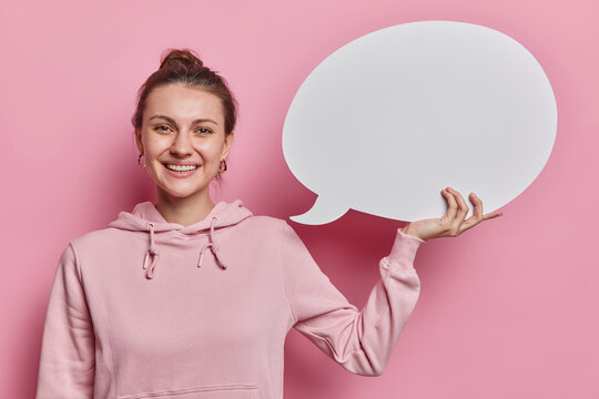 Young pretty adult woman holds white speech bubble over pink background dressed in casual hoodie suggests to write your text promotional content or idea there. Blank word box free space. Mock up.