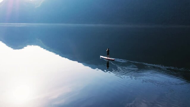 Epic and cinematic serene aerial shot of single paddle board move through still fjord water in Norway. Stand up paddle board in amazing scenery. Drone shot of paddle board in fjord