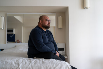 Frustrated overweight man sits on bed sadly looking to window feeling depression from difficult...
