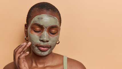 Headshot of dark skinned female model applies facial beauty mask to reduce pores keeps eyes closed enjoys skin care treatments stands bare shoulders isolated over brown background copy space for text - obrazy, fototapety, plakaty