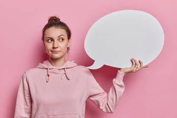 Thoughtful pretty young woman with dark hair gathered in bun holds abstract cloud bubble for thoughts and words dressed in casual sweatshirt isolated over pink background. Communication concept