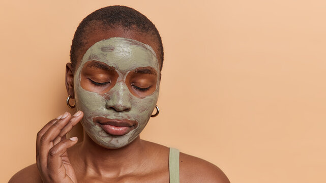Headshot of dark skinned female model applies facial beauty mask to reduce pores keeps eyes closed enjoys skin care treatments stands bare shoulders isolated over brown background copy space for text