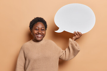 Young cheerful African woman with dark hair holds blank empty white speech bubble for your design smiles broadly dressed in casual knitted jumper isolated on brown background. News dialog announcement