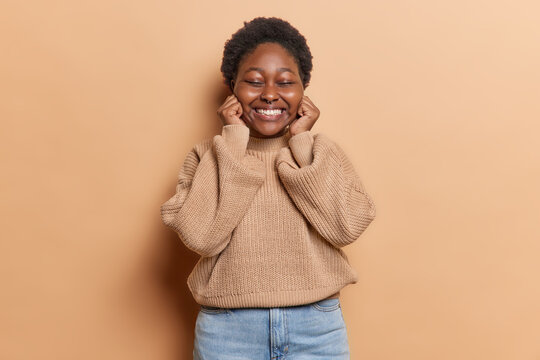 Positive sincere human emotions concept. Plump overjoyed woman keeps hands on cheeks has eyes closed smiles broadly dressed in loose knitted sweater and jeans isolated over brown background.