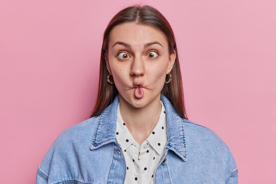 Portrait of funny brunette European woman makes ridiculous childish grimace has fish lips playful comical expression has fun foolishes around wears denim jacket isolated over pink background.