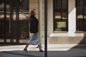 Beautiful brunette woman walks street city near buildings, wears luxurious attire clothes shiny maxi skirt with sequins, black sneakers and Tshirt. Light and shadow - obrazy, fototapety, plakaty