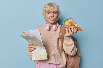 Female traveler holds paper map searches for route eats sandwich purses lips has dimpes in cheeks...