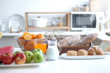 Variety of fresh fruits and bread on table in bright white kitchen, healthy lifestyle concept, glass of milk and orange Juice