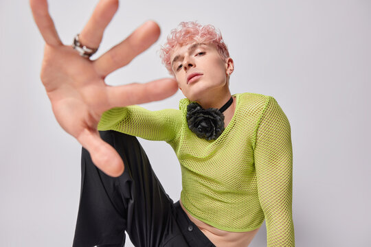 Serious self confident androgynous pink haired man keeps hand towards camera wears green net jumper trousers poses against white background. Pink haired gay looks through fingers. Homosexuality - Powered by Adobe