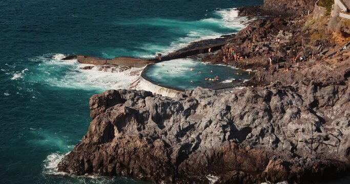 Stormy ocean waves crash on rocky shore of natural swimming pool in Tenerife. Charco de Isla Cangrejo. People sunbathing on volcanic beach.