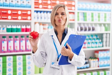 Young woman working at pharmacy drugstore holding heart looking at the camera blowing a kiss being...